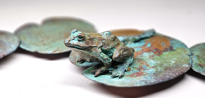 K&ouml;nnte beinhalten: Eine kleine gr&uuml;ne Froschskulptur, die auf einem kupferfarbenen Seerosenblatt sitzt. Der Frosch ist mit strukturierter Haut detailliert und hat ein realistisches Aussehen.