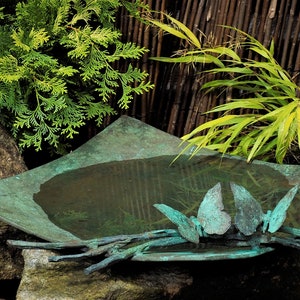 May include: A square, teal-colored metal bird bath with a stylized butterfly design. The bird bath is filled with water and sits on a rock in a garden setting.
