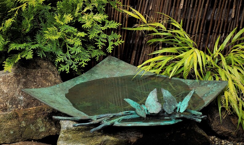 May include: A square, teal-colored metal bird bath with a stylized butterfly design. The bird bath is filled with water and sits on a rock in a garden setting.