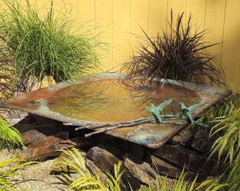 Puede incluir: Un baño de pájaros de cobre con una cuenca poco profunda llena de agua. El baño de pájaros está rodeado de rocas y plantas verdes. Dos colibríes de bronce están posados en el borde de la cuenca.