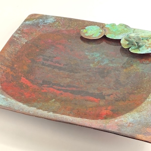 K&ouml;nnte beinhalten: Ein quadratisches Kupfertablett mit einer erh&ouml;hten, abgerundeten Mitte und einer marmorierten, mehrfarbigen Oberfl&auml;che. Das Tablett hat eine gr&uuml;ne Patina und ein dekoratives Design aus stilisierten Seerosenbl&auml;ttern.
