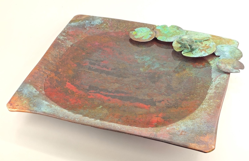 K&ouml;nnte beinhalten: Ein quadratisches Kupfertablett mit einer erh&ouml;hten, abgerundeten Mitte und einer marmorierten, mehrfarbigen Oberfl&auml;che. Das Tablett hat eine gr&uuml;ne Patina und ein dekoratives Design aus stilisierten Seerosenbl&auml;ttern.