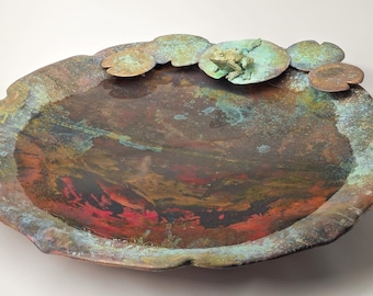 Puede incluir: Un plato de cobre con una superficie texturizada y moteada en tonos marrones, rojos y verdes. El plato tiene forma de estanque con nenúfares y una rana en el borde.
