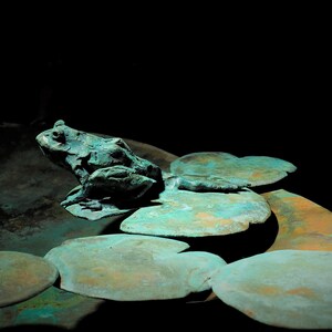 K&ouml;nnte beinhalten: Eine gr&uuml;ne Froschskulptur, die auf einem Bett aus Seerosenbl&auml;ttern sitzt, alles aus Metall mit einer Gr&uuml;nspan-Oberfl&auml;che.