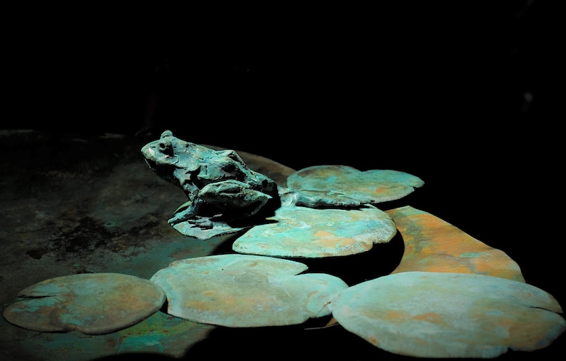 K&ouml;nnte beinhalten: Eine gr&uuml;ne Froschskulptur, die auf einem Bett aus Seerosenbl&auml;ttern sitzt, alles aus Metall mit einer Gr&uuml;nspan-Oberfl&auml;che.