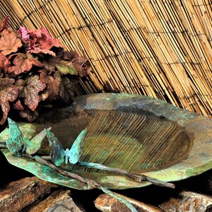 May include: A rustic, green metal bird bath with a shallow basin and a decorative metal butterfly design. The bird bath is sitting on a pile of brown rocks in front of a bamboo fence.