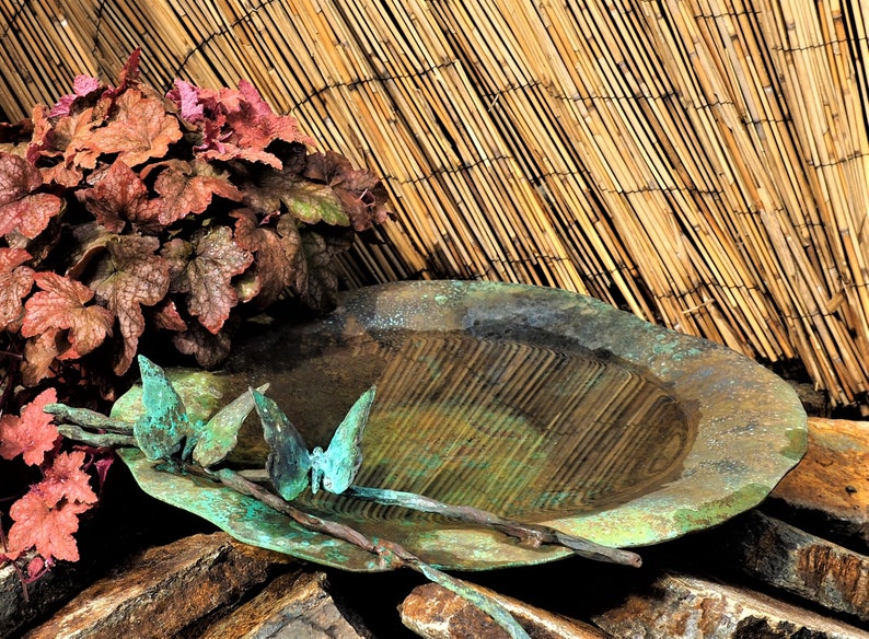 May include: A rustic, green metal bird bath with a shallow basin and a decorative metal butterfly design. The bird bath is sitting on a pile of brown rocks in front of a bamboo fence.