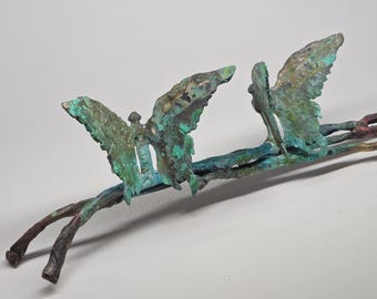 Puede incluir: Escultura de bronce con dos mariposas estilizadas posadas en una rama. Las mariposas tienen alas texturizadas con pátina de verdín, mientras que la rama es una mezcla de marrón y verde. La escultura está sobre un fondo liso.