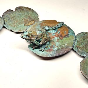 K&ouml;nnte beinhalten: Eine Bronzeskulptur eines Frosches, der auf einem Seerosenblatt sitzt. Die Skulptur besteht aus mehreren Teilen, die jeweils eine andere Patina haben. Der Frosch ist detailliert und realistisch, mit strukturierter Haut und einer lebensechten Pose.