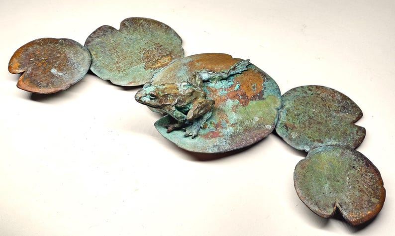 K&ouml;nnte beinhalten: Eine Bronzeskulptur eines Frosches, der auf einem Seerosenblatt sitzt. Die Skulptur besteht aus mehreren Teilen, die jeweils eine andere Patina haben. Der Frosch ist detailliert und realistisch, mit strukturierter Haut und einer lebensechten Pose.