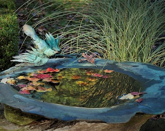 May include: A blue metal bird sculpture perched on the edge of a stone birdbath. The birdbath is filled with water and has fallen leaves floating on the surface. The birdbath is surrounded by green grass and foliage.