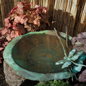 May include: A copper bird bath with a dragonfly sculpture and a wooden stick. The bird bath is surrounded by red and green foliage.