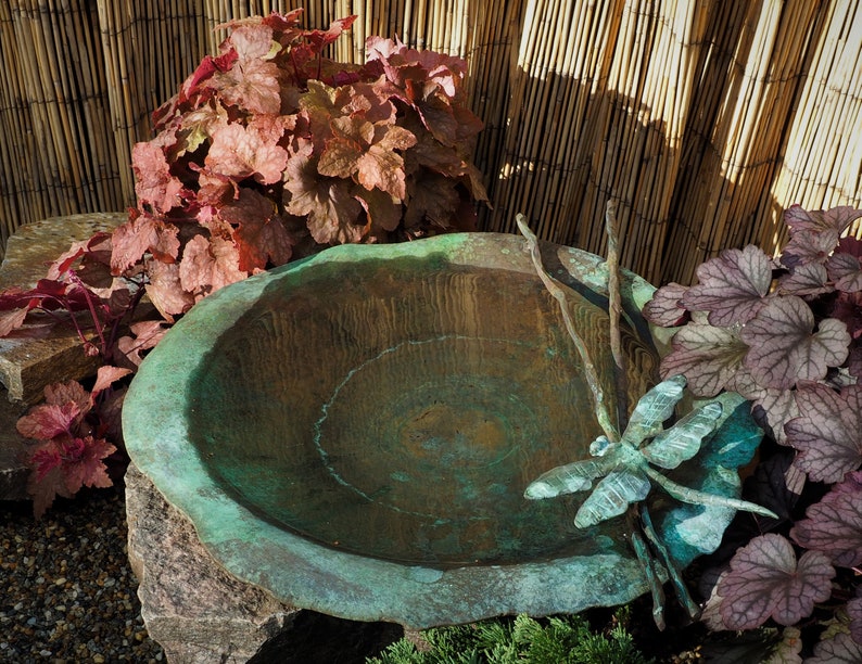 May include: A copper bird bath with a dragonfly sculpture and a wooden stick. The bird bath is surrounded by red and green foliage.