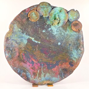 K&ouml;nnte beinhalten: Eine runde Wandskulptur aus Kupfer mit einer strukturierten, marmorierten Oberfl&auml;che in Braun-, Gr&uuml;n-, Blau- und Rosat&ouml;nen. Die Skulptur zeigt mehrere erhabene, abgerundete Formen, die an Seerosenbl&auml;tter erinnern, wobei ein Seerosenblatt eine kleine Froschfigur tr&auml;gt.