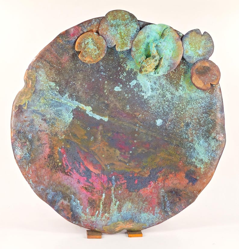 K&ouml;nnte beinhalten: Eine runde Wandskulptur aus Kupfer mit einer strukturierten, marmorierten Oberfl&auml;che in Braun-, Gr&uuml;n-, Blau- und Rosat&ouml;nen. Die Skulptur zeigt mehrere erhabene, abgerundete Formen, die an Seerosenbl&auml;tter erinnern, wobei ein Seerosenblatt eine kleine Froschfigur tr&auml;gt.