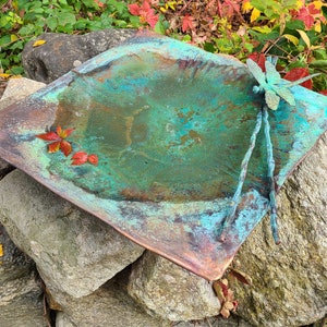 May include: A copper bird bath with a dragonfly sculpture on top. The bird bath is green and blue with a rustic patina. It is sitting on a pile of rocks.