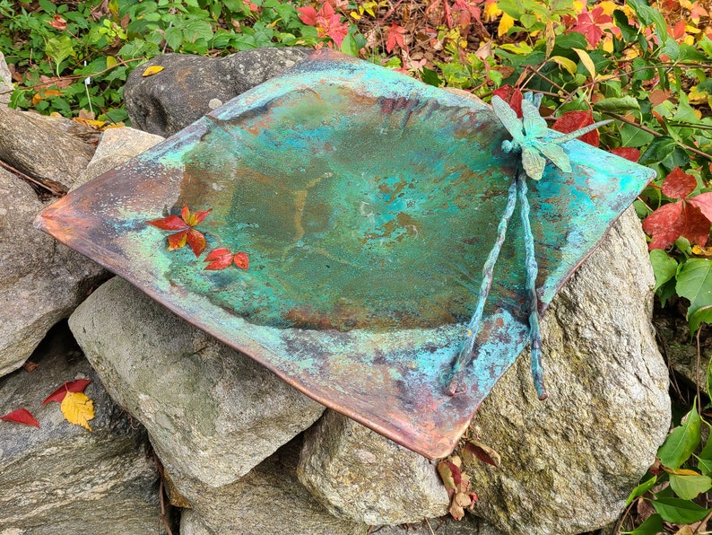 May include: A copper bird bath with a dragonfly sculpture on top. The bird bath is green and blue with a rustic patina. It is sitting on a pile of rocks.