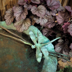 May include: A green metal dragonfly sculpture with intricate details sits on a copper-colored dish. The dragonfly is perched on a branch with its wings spread wide. The dish is surrounded by purple-hued leaves.