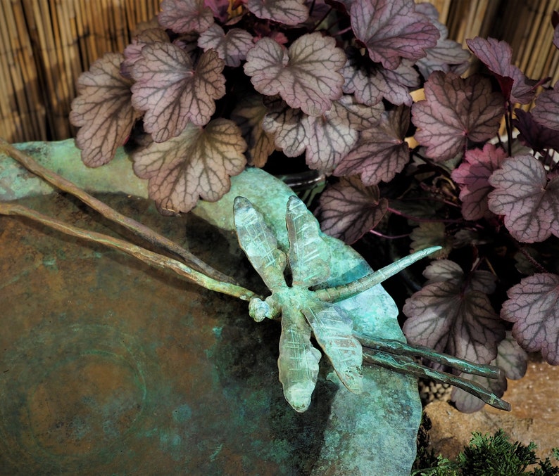 May include: A green metal dragonfly sculpture with intricate details sits on a copper-colored dish. The dragonfly is perched on a branch with its wings spread wide. The dish is surrounded by purple-hued leaves.