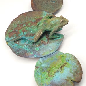 K&ouml;nnte beinhalten: Eine gr&uuml;ne Froschskulptur, die auf einer gr&uuml;nen und braunen Seerosenblatt-Skulptur sitzt. Das Seerosenblatt ist aus Metall und hat eine strukturierte Oberfl&auml;che.