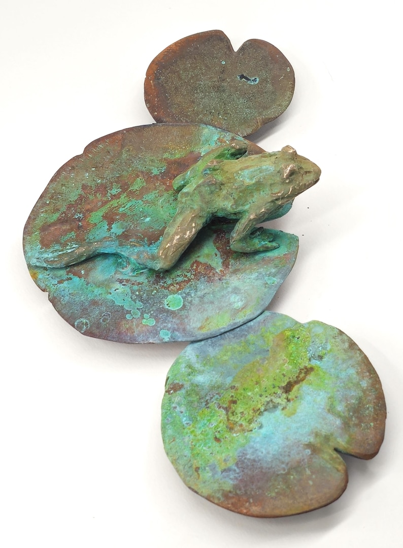 K&ouml;nnte beinhalten: Eine gr&uuml;ne Froschskulptur, die auf einer gr&uuml;nen und braunen Seerosenblatt-Skulptur sitzt. Das Seerosenblatt ist aus Metall und hat eine strukturierte Oberfl&auml;che.