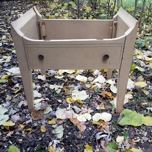 May include: A beige wooden table with a single drawer and rounded edges. The table is unfinished and has a rustic look. The table is sitting on a bed of fallen leaves.