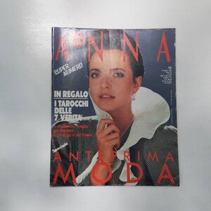 May include: A vintage magazine cover featuring a woman with dark hair and a white ruffled collar. The magazine title is "ANNA" in red letters, with additional text in Italian. The cover has a blue background and a checkered pattern.