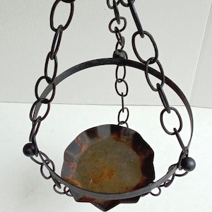 May include: A hanging metal candle holder with a circular design. The holder is suspended by chains and features a round, scalloped tray. The metal has a dark, aged finish, suggesting a vintage or rustic aesthetic.