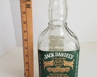 May include: A clear glass bottle with a green label that reads "Jack Daniel's Old Time Distillery No. 7 Brand Sour Mash Tennessee Whiskey". The label also includes the text "Quality" and "Jack Daniel Distillery, Lynchburg, Tennessee, Est. 1866".