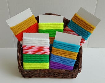 May include: A brown woven wicker basket containing five rectangular white cardboard cards wrapped with multi-colored yarn. The yarn colors include yellow, orange, neon green, white, pink, red, blue, purple, and turquoise in various striped patterns.