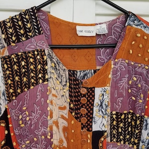 May include: A sleeveless patchwork dress with a brown, purple, and white floral print. The dress has a scoop neck and a button closure at the front. The label reads "Ann Ashley".
