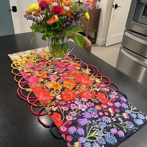 May include: A rectangular fabric table runner featuring a vibrant floral print with a gradient of colors from yellow and orange to pink, red, and purple. The runner has decorative fabric loops along both sides and a glass vase of flowers.