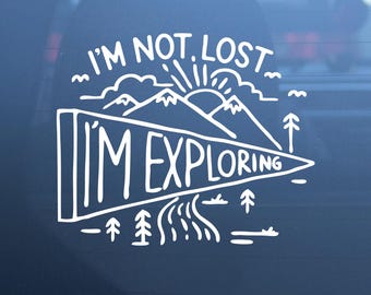 May include: A white vinyl decal featuring a mountain landscape with a sun, trees, and a winding river. The text reads I'M NOT LOST I'M EXPLORING, with the words I'M EXPLORING contained within a large triangular pennant shape.