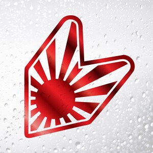 May include: A red and white graphic sticker featuring a rising sun design within a stylized arrow shape. The sun has multiple rays emanating from a central red circle. The sticker has a glossy finish.