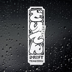 以下が含まれることがあります： 黒い枠の中に、日本語の「ドリフト」という単語をスタイリッシュにデザインした白いステッカー。ステッカーの下には「ドリフト天国」という文字が書かれている。