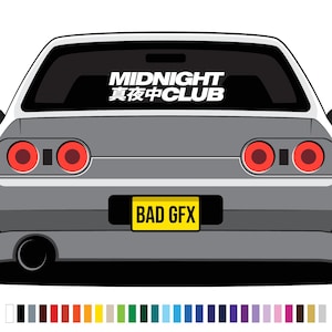 May include: A grey car's rear, featuring red circular taillights and a black window sticker with the white text "MIDNIGHT 真夜中CLUB". The yellow license plate displays "BAD GFX" in black.