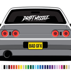 以下が含まれることがあります： 後部窓に白い「DRIFT MISSILE」のステッカーが貼られた灰色の車。黄色いナンバープレートには「BAD GFX」と書いてある。