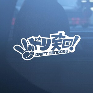 May include: A white vinyl decal sticker featuring a hand making a peace sign gesture next to Japanese characters and the text DRIFT TENGOKU in a bold, stylised font. The design is applied to a dark, smooth, reflective surface.