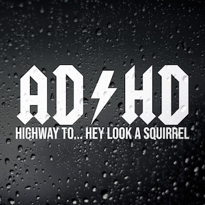 Peut inclure: Autocollant en vinyle blanc avec le texte "ADHD" dans une police de caractères stylisée et audacieuse. Le texte est suivi de "Highway to... Hey Look A Squirrel" dans une police plus petite.
