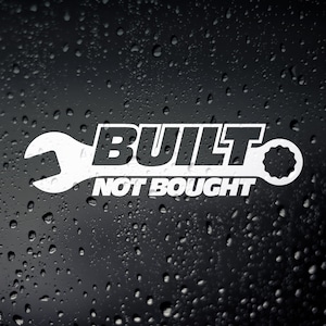 Peut inclure: Graphique blanc avec le texte "BUILT NOT BOUGHT" en gras, police sans empattement. Le mot "BUILT" est intégré à un design de clé. Le fond est une surface sombre avec des gouttelettes d'eau.