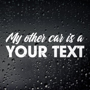 以下が含まれることがあります： 「My other car is a YOUR TEXT」というテキストが筆記体フォントで書かれた白いビニールデカール。デカールは水滴のある黒い背景にあります。