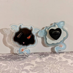 May include: Two clear acrylic pins featuring black and white cartoon characters. The black character has red eyes and a tongue sticking out. The white character has a black heart-shaped pin back. Both characters have blue wings and tails.