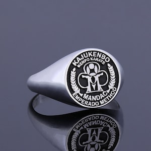 May include: A silver signet ring with a black enamel design. The design features the words "Kajukenbo Kenpo Karate" and "Emperorado Method" surrounding a logo with a stylized "M" and a martial arts figure.