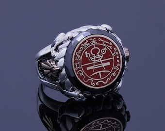May include: A silver ring with a red stone featuring a silver sigil. The ring has a detailed design with a black background.
