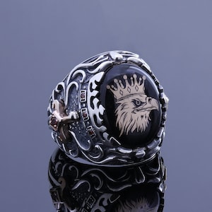 May include: A silver ring with an ornate design featuring a black onyx stone with a gold eagle wearing a crown. The ring is set against a gray background.