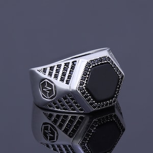 May include: A silver ring with a black hexagonal stone set in the center. The ring is decorated with small black stones around the stone and the band.