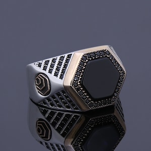 May include: A silver ring with a black onyx stone set in a hexagonal shape. The ring is accented with small black stones around the stone and the band.