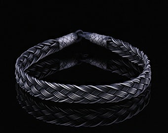 May include: A silver braided bracelet with a woven design. The bracelet is on a black background.