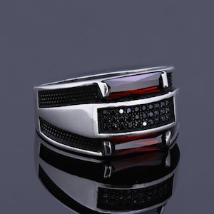 Op de afbeelding: Een zilveren ring met een ontwerp van zwarte en rode stenen. De ring heeft een getextureerde band en een rechthoekige rode steen die in een zilveren bezel is gezet. De steen is omgeven door kleinere zwarte stenen.