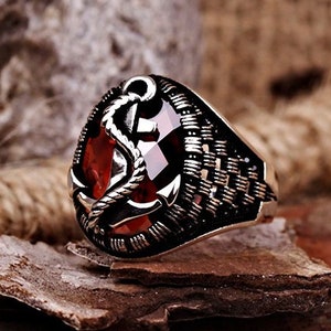 May include: A silver ring with a red gemstone and an anchor design. The ring is set on a rustic background.